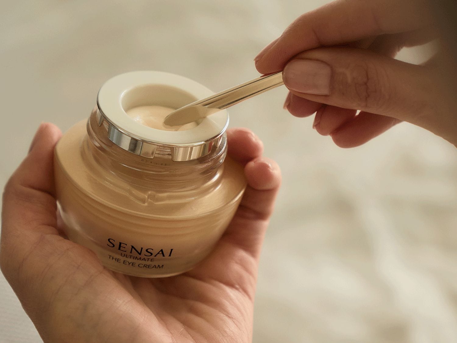 Detailní pohled na Sensai Ultimate The Eye Cream v béžovém balení, nabíraný zlatou špachtlí, slibující luxusní péči o oční okolí.