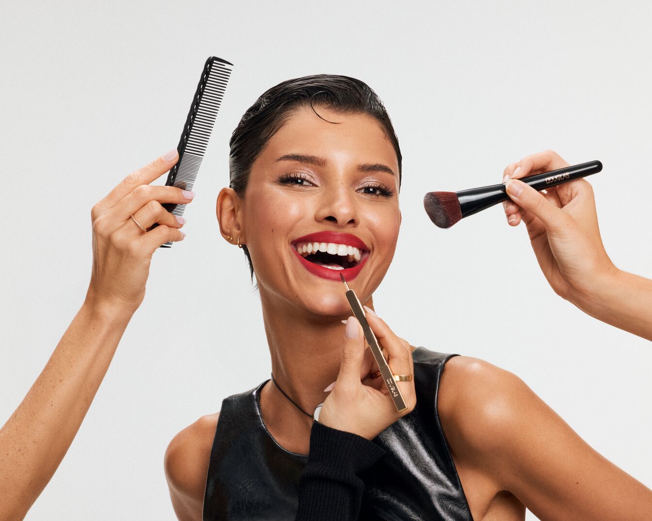 Eine Frau mit glänzenden, kurzen Haaren und rotem Lippenstift lächelt, während sie von einem Kamm, einem Pinsel und einem Lippenstift umgeben ist.