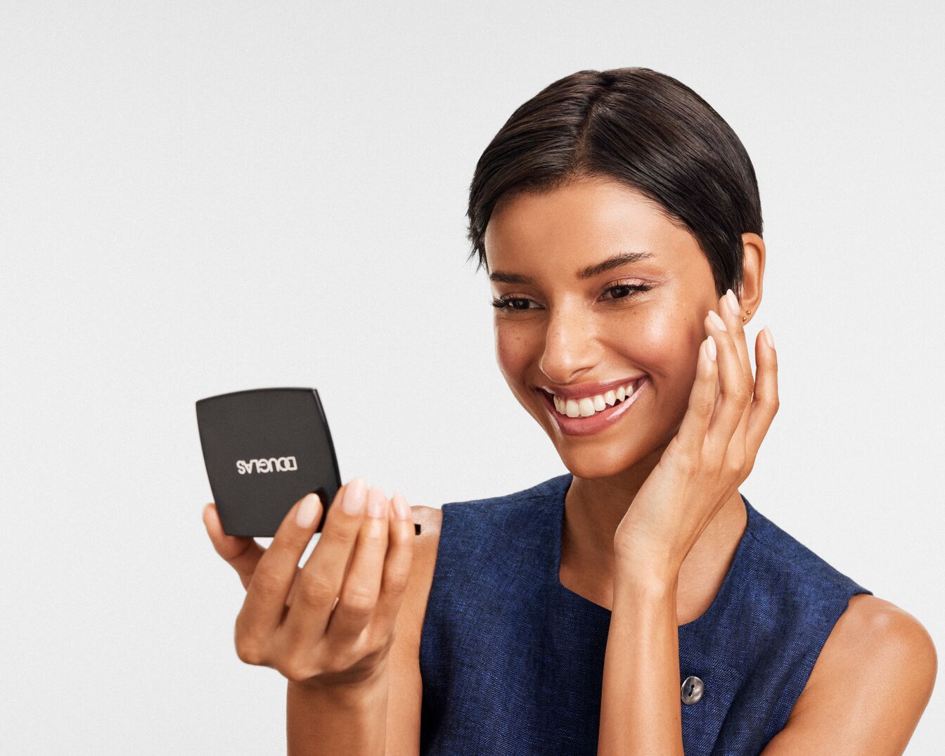 Eine lächelnde Frau mit kurzem, dunklem Haar betrachtet ihr strahlendes Make-up in einem schwarzen Douglas-Kompaktspiegel.