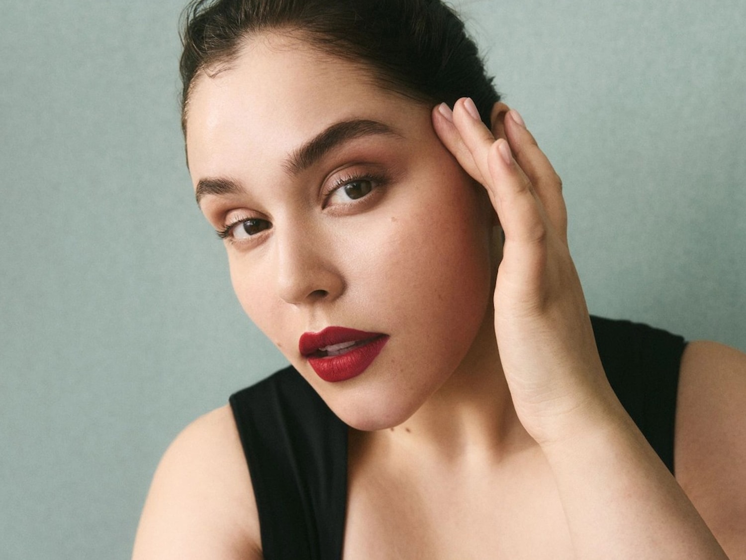 Portrait d'une femme avec un rouge à lèvres rouge foncé et un fard à paupières marron, touchant son visage avec sa main.