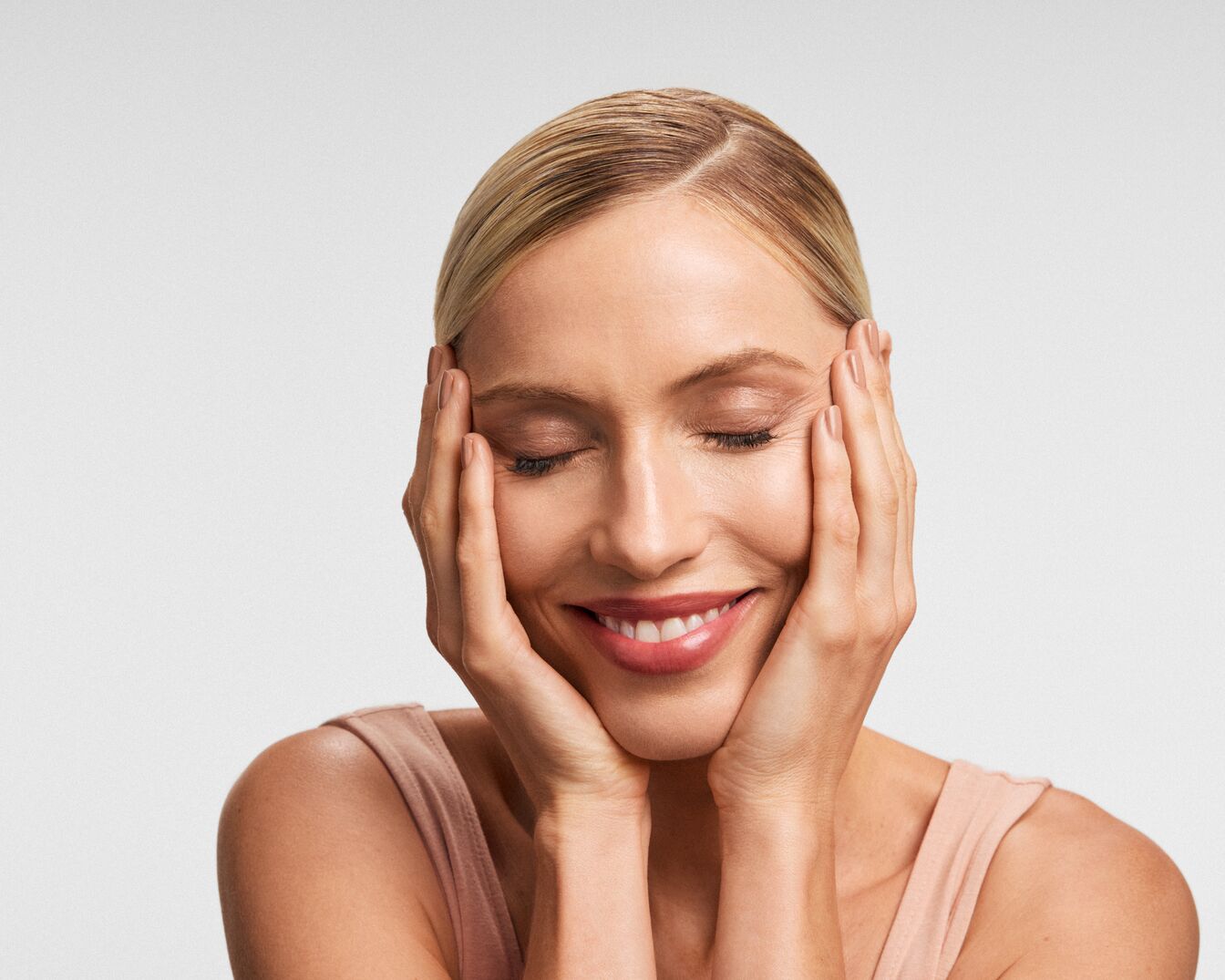 Eine blonde Frau mit geschlossenen Augen und einem sanften Lächeln hält ihre Hände an den Wangen.