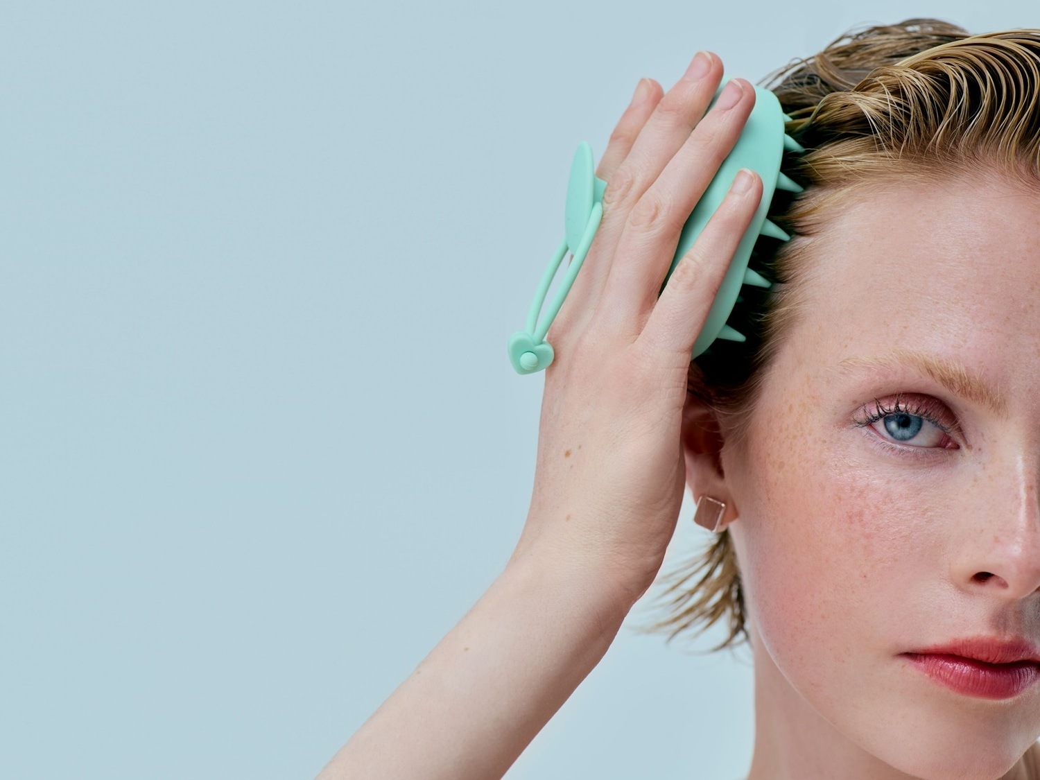 Eine Frau mit hellblonden, nassen Haaren massiert ihre Kopfhaut mit einer türkisfarbenen Bürste.