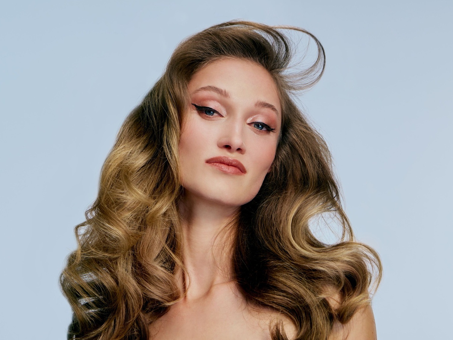 Porträt einer Frau mit gewelltem, braunem Haar, zartem rosa Lidschatten und einem präzisen schwarzen Eyeliner, der ihren Blick betont.