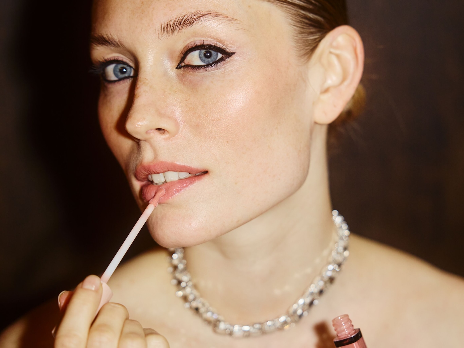 Una donna con eyeliner nero e labbra rosa lucide applica un lucidalabbra rosa chiaro con un applicatore, indossando una collana di strass.