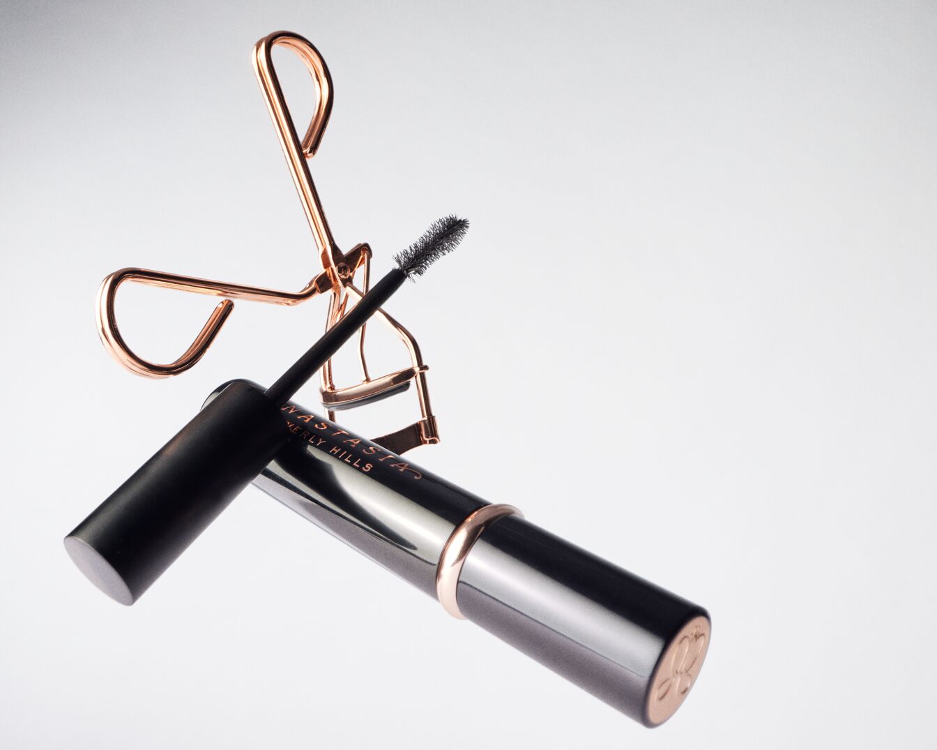 Un piegaciglia color oro rosa e un mascara nero con il marchio Anastasia Beverly Hills sono mostrati su uno sfondo bianco, con lo scovolino del mascara che sporge dal tubetto.