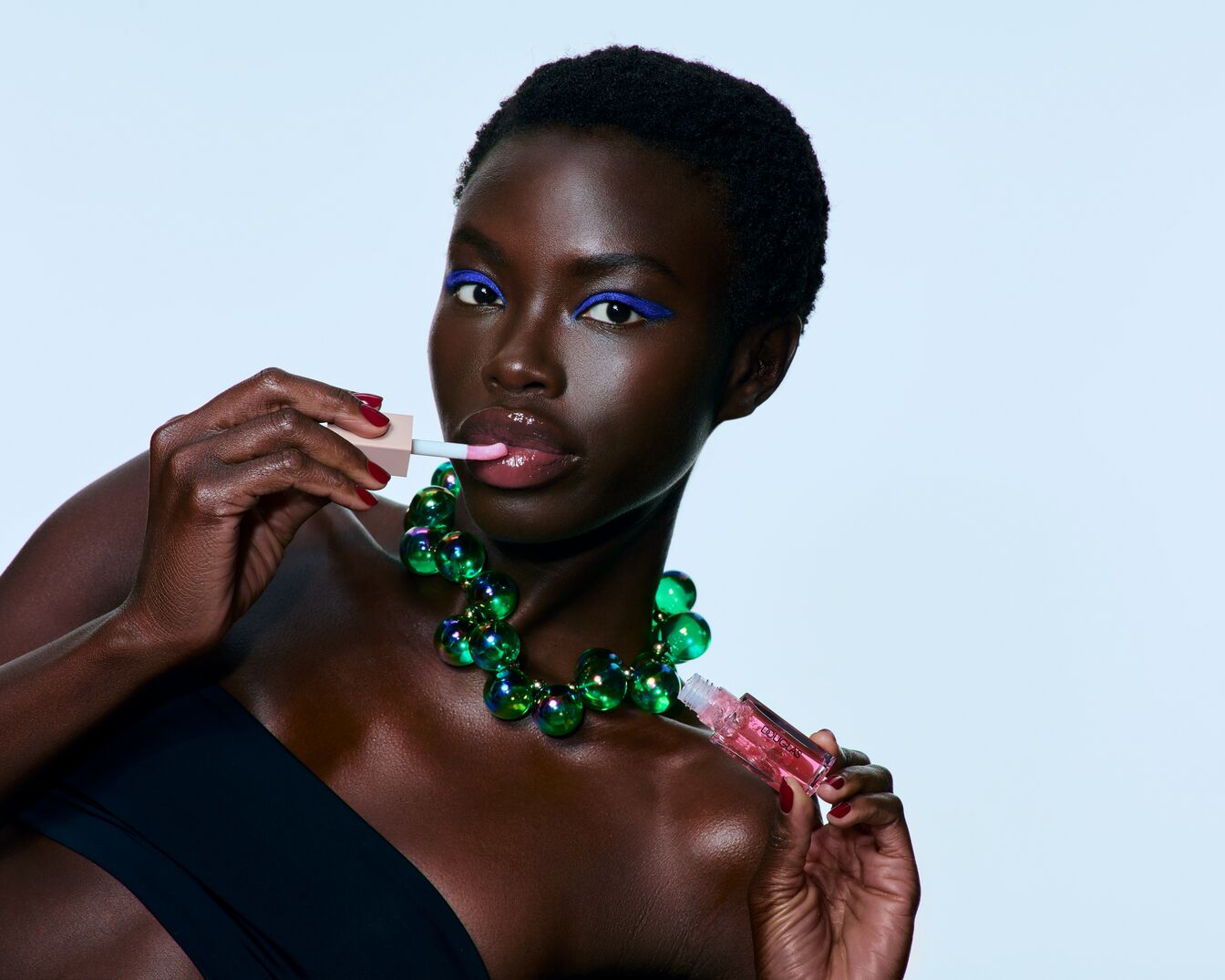 Eine Frau mit Schwarzer Haut trägt einen grünen Kugelkette und blauen Eyeliner, während sie einen rosa Lipgloss mit einem beigen Applikator aufträgt.