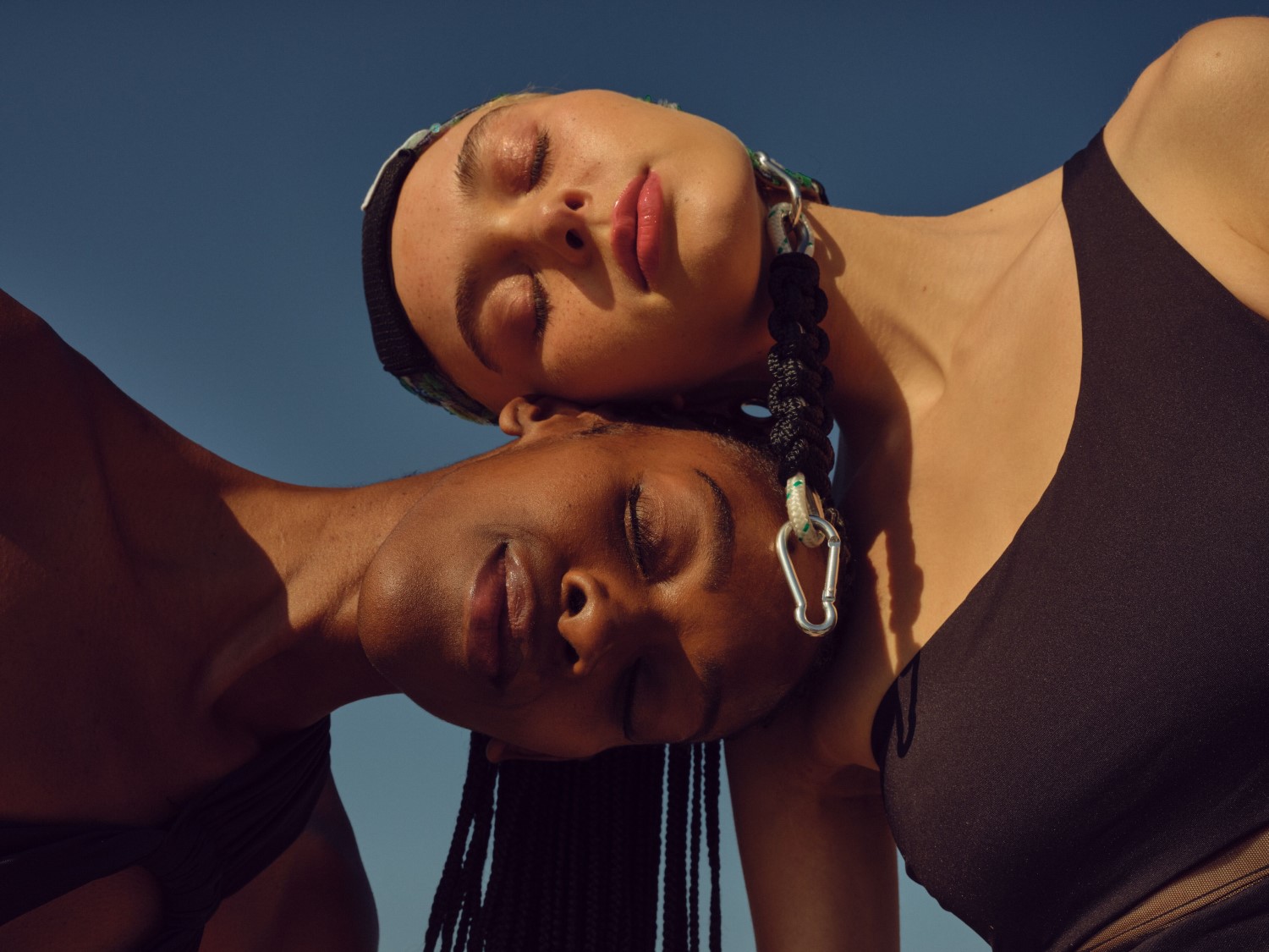 Dve ženy s tmavými vlasmi a čiernymi plavkami sa opierajú hlavami o seba na pozadí modrej oblohy, pričom jedna má na hlave šatku a náušnice.