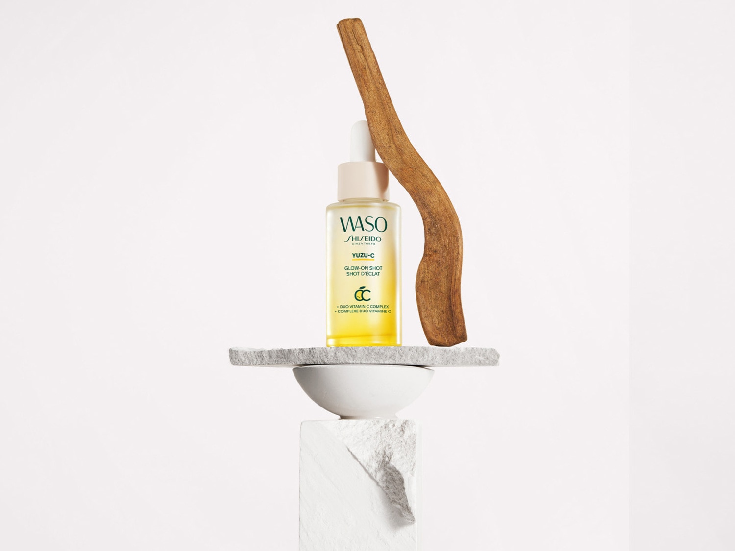 Een flesje Shiseido WASO Yuzu-C Glow-On Shot staat op een stenen sokkel, met een houten tak ernaast, wat de natuurlijke ingrediënten benadrukt.