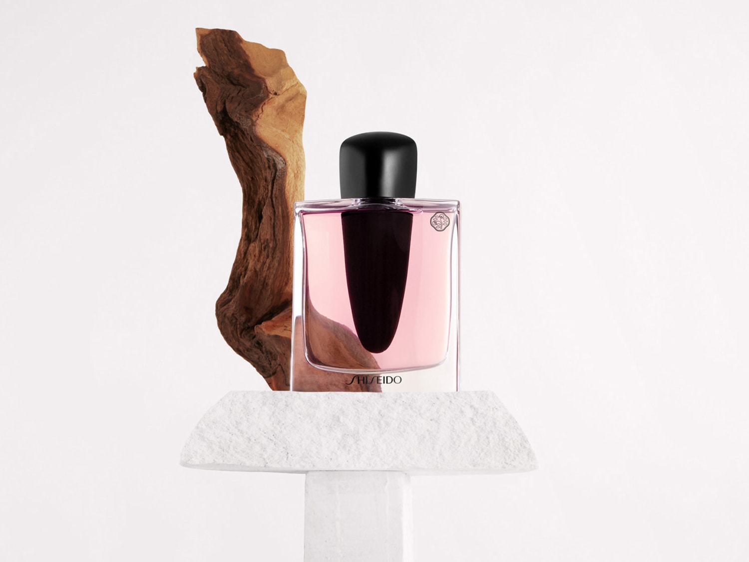Een rechthoekige Shiseido parfumfles met een zwarte dop, gevuld met roze vloeistof, staat op een witte stenen sokkel naast een stuk drijfhout.