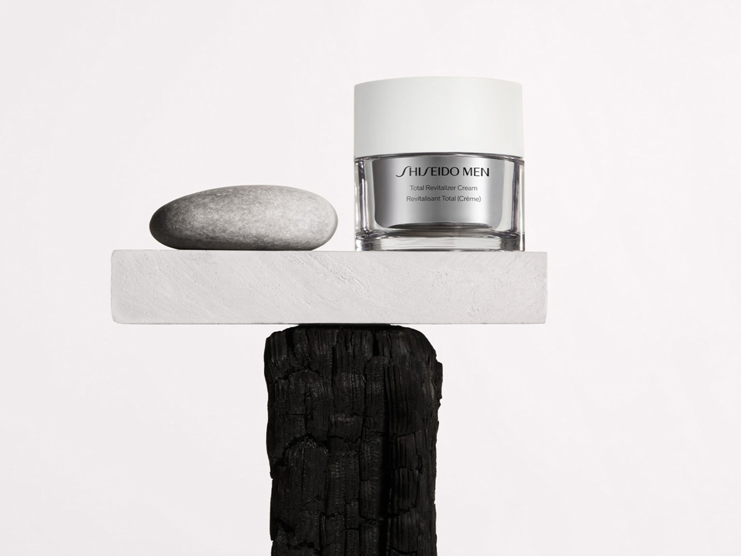 Shiseido Men Total Revitalizer Cream in een heldere pot met zilveren accenten, gepresenteerd op een rustieke houten sokkel naast een gladde, grijze steen.