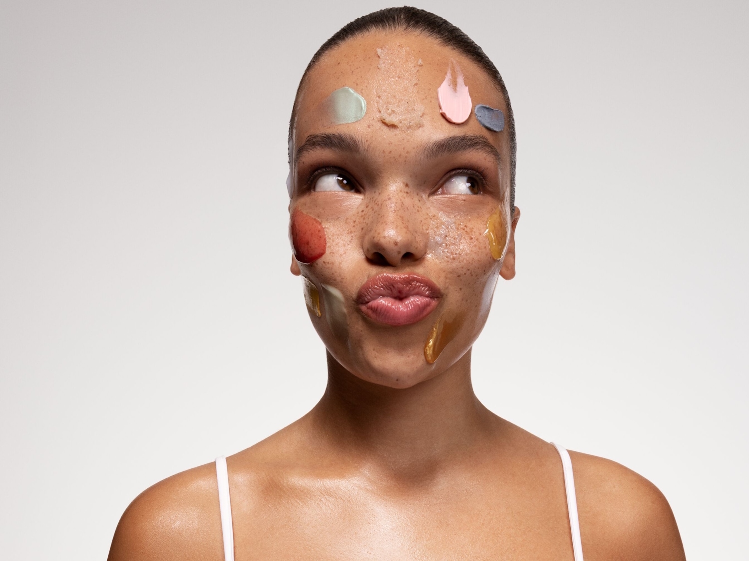 Eine Frau mit Sommersprossen hat verschiedene farbige Cremes auf ihrem Gesicht aufgetragen, darunter Beige, Rosa, Grün, Rot, Gelb und Grau, während sie mit zusammengepressten Lippen zur Seite schaut.