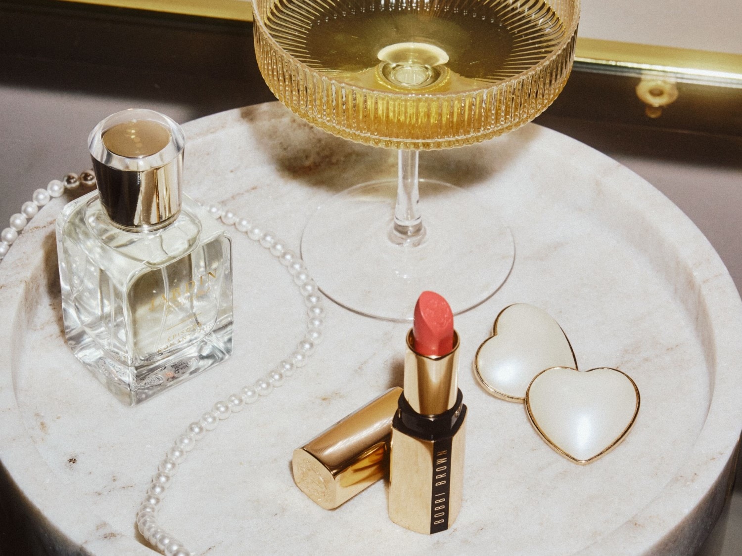 Na mramorovém podnose je parfém v čtvercové lahvičce, rtěnka Bobbi Brown ve zlatém obalu, sklenice šampaňského a perlový náhrdelník.
