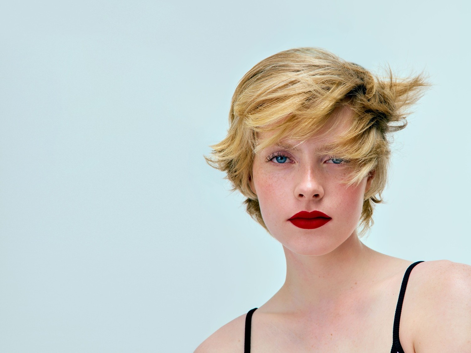 Portret van een vrouw met blond, kort haar, blauwe ogen en rode lippenstift, tegen een lichtblauwe achtergrond.