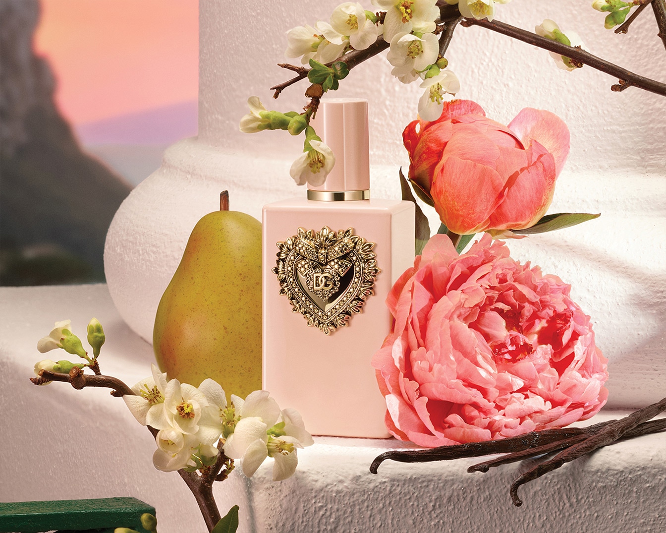 Un flacone di profumo rosa chiaro Dolce&Gabbana con un ornamento dorato a forma di cuore, affiancato da una pera verde, fiori bianchi e peonie rosa.