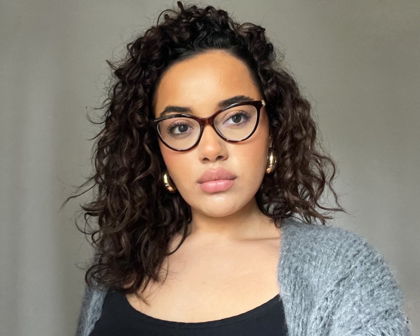Porträt einer jungen Frau mit lockigem Haar, Brille und goldenen Ohrringen, die einen grauen Cardigan trägt.