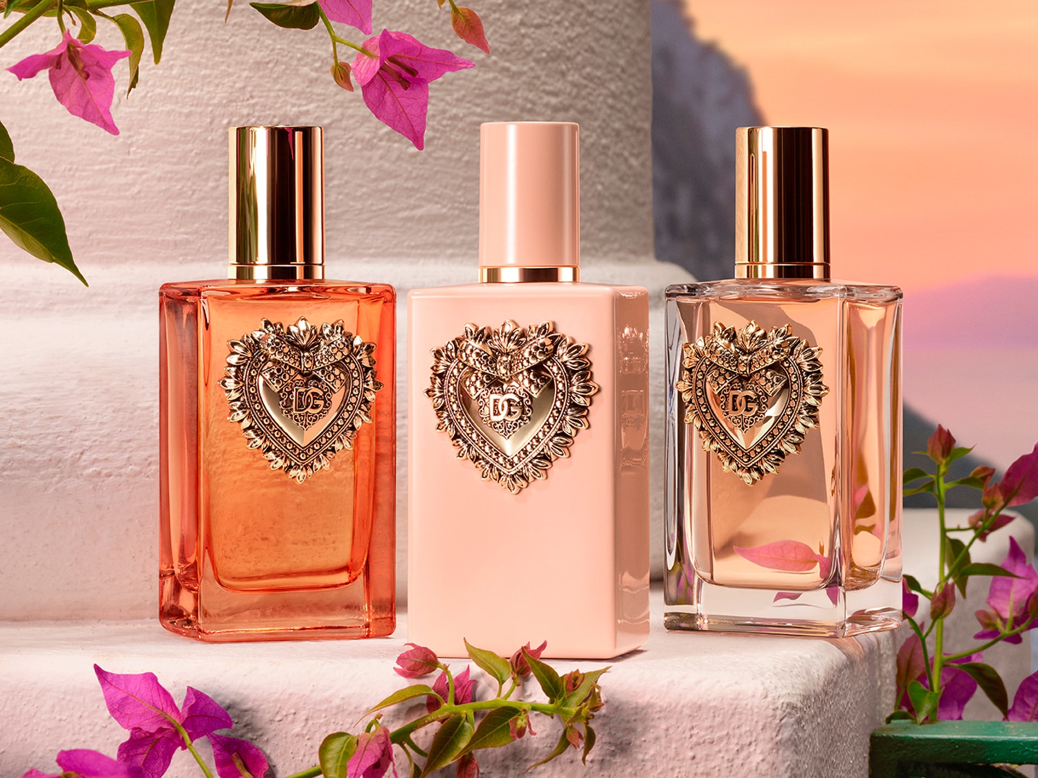 Tres frascos de perfume Dolce&Gabbana con forma rectangular y tapones dorados o color melocotón, adornados con un corazón dorado con las iniciales DG, sobre un fondo floral.