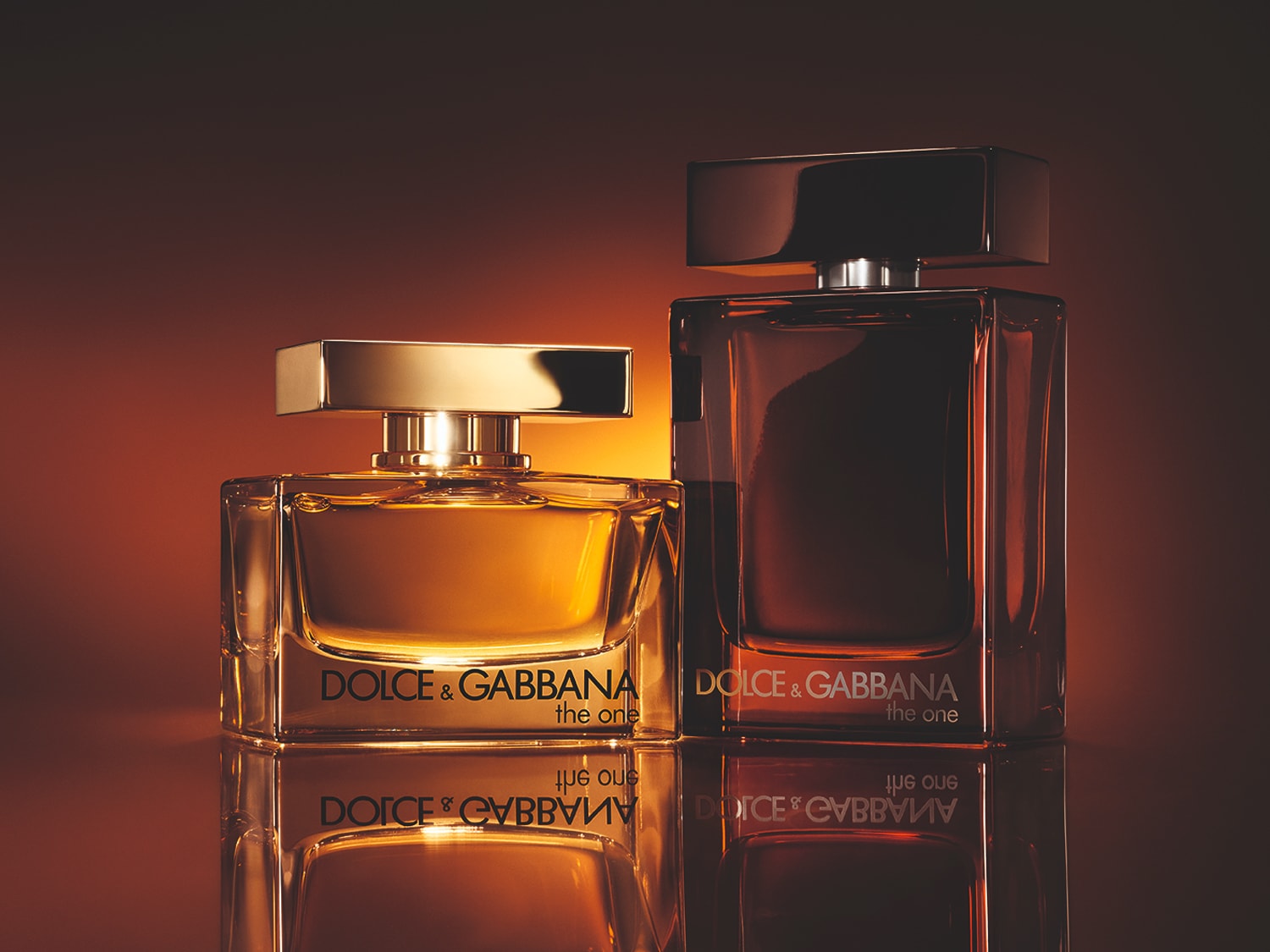 Dos frascos de perfume Dolce & Gabbana The One, uno dorado y otro marrón oscuro, sobre una superficie reflectante con un fondo cálido y difuminado.