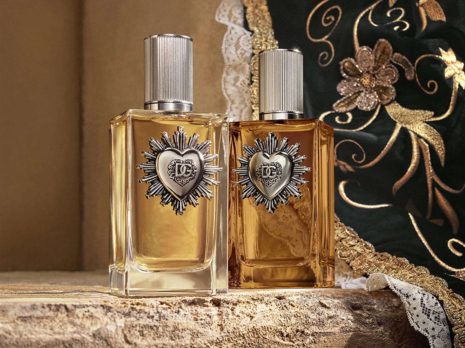 Dos frascos de perfume Dolce&Gabbana con forma rectangular y tapón plateado, adornados con un corazón metálico con el logo de DG, uno con líquido dorado claro y otro con líquido dorado oscuro.