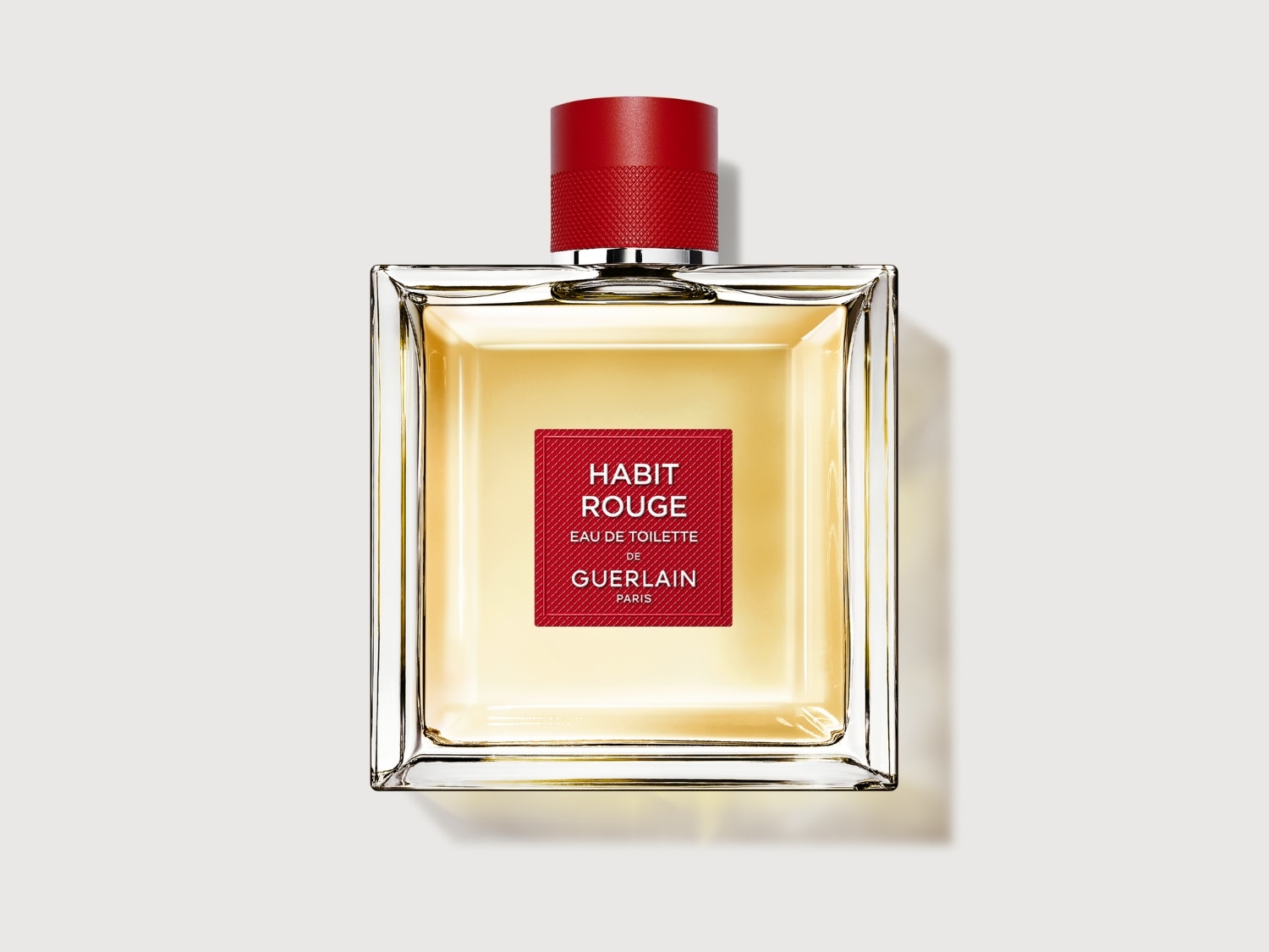 El frasco cuadrado de cristal transparente contiene el perfume Habit Rouge Eau de Toilette de Guerlain, con una etiqueta roja y un tapón rojo texturizado.