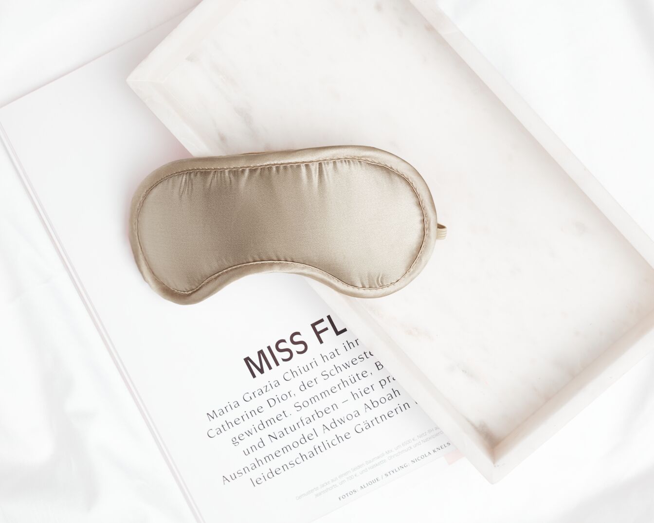 Nahaufnahme einer goldenen Schlafmaske aus Seide, die auf einer Zeitschrift mit dem Titel MISS FL und einem Marmortablett auf einem weißen Tuch liegt.