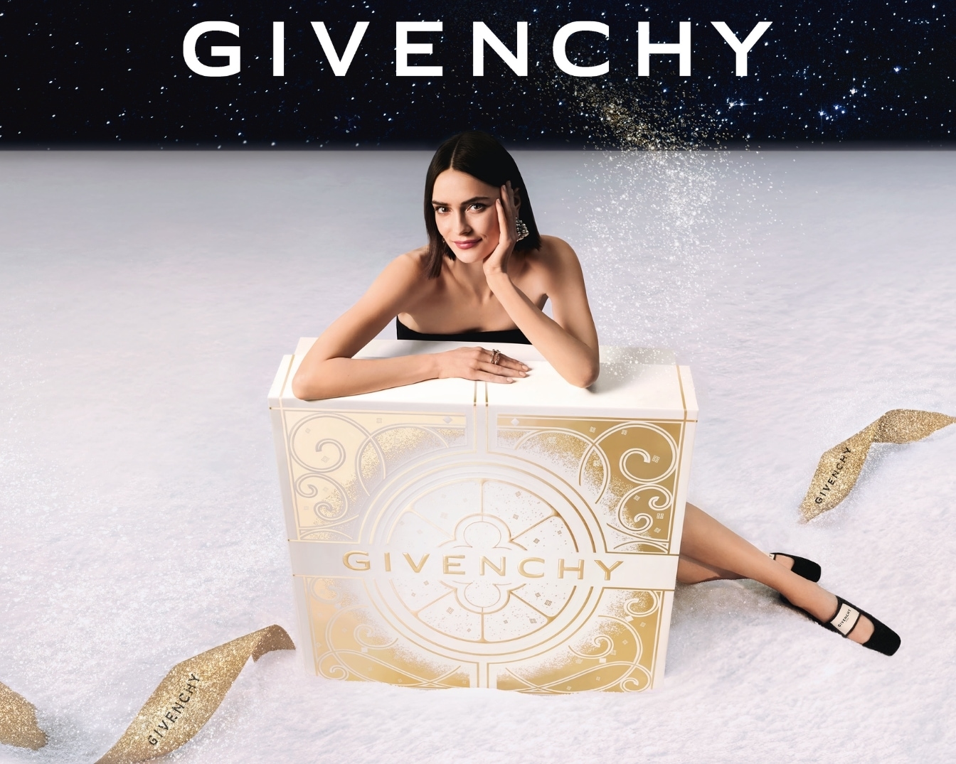 Eine Frau posiert hinter einer weißen und goldenen Givenchy-Box vor einem sternenklaren Hintergrund, umgeben von goldenem Glitzer.