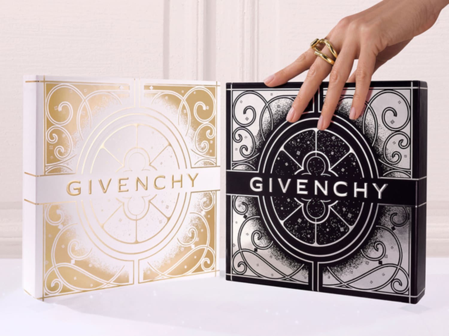 Zwei Givenchy-Boxen, eine in Weiß mit goldenen Verzierungen und die andere in Schwarz mit silbernen Details, werden von einer Hand mit goldenen Ringen berührt.