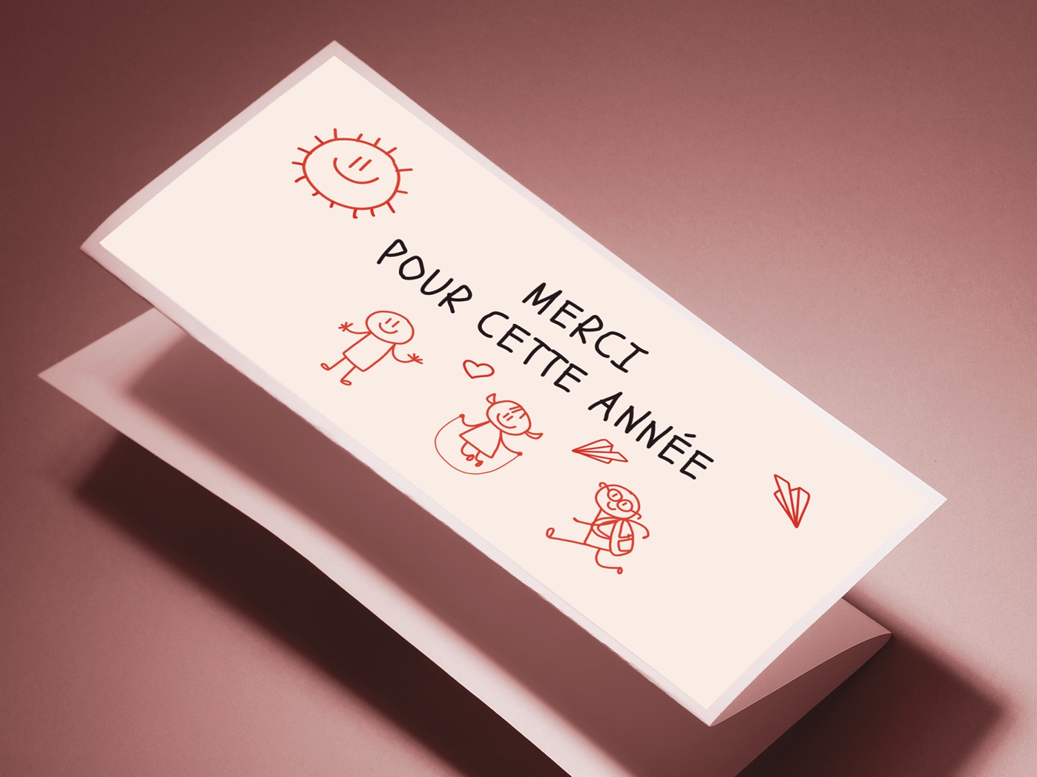 Une carte de remerciement blanche avec l'inscription "MERCI POUR CETTE ANNÉE" en rouge, accompagnée de dessins d'enfants et d'un soleil souriant, posée sur un fond rose.