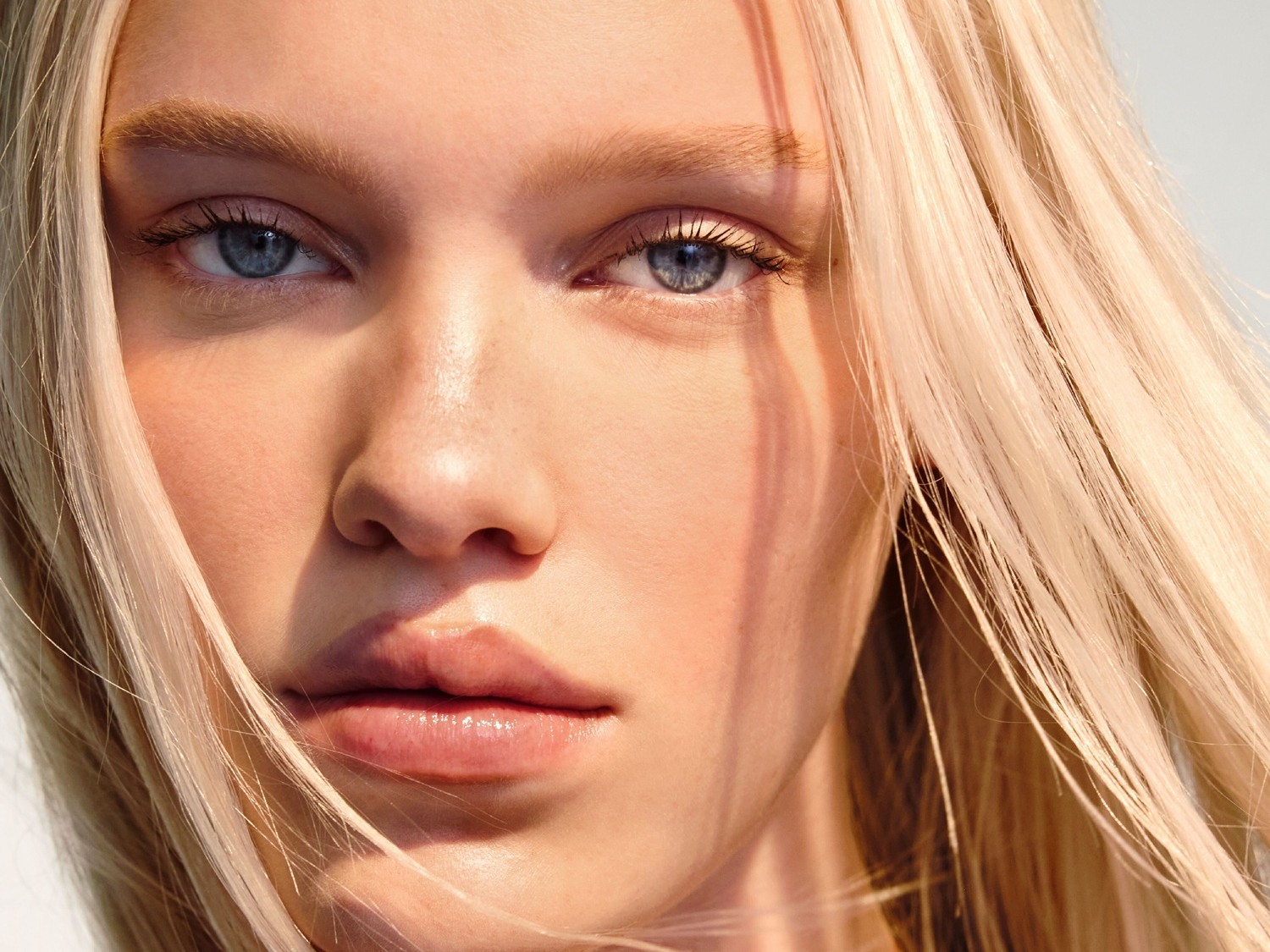 Nahaufnahme eines hellhäutigen Models mit blonden Haaren und blauen Augen, das einen natürlichen, strahlenden Teint und glänzende Lippen zeigt.