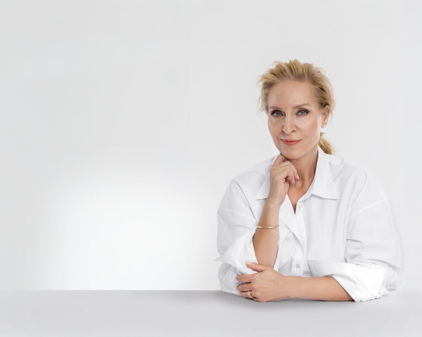 Eine blonde Frau mit einem weißen Hemd sitzt an einem Tisch, stützt ihr Kinn auf die Hand und blickt direkt in die Kamera.