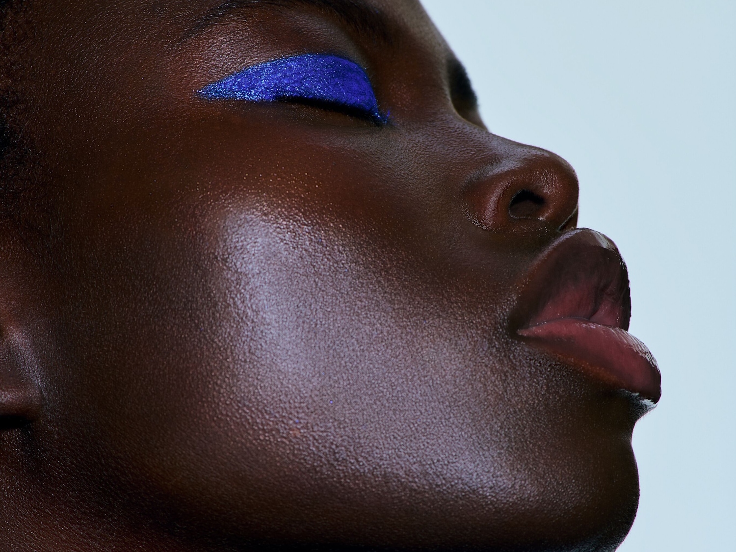 Nahaufnahme eines dunklen Hauttons mit schimmerndem blauem Eyeliner auf dem Augenlid und glänzenden Lippen.