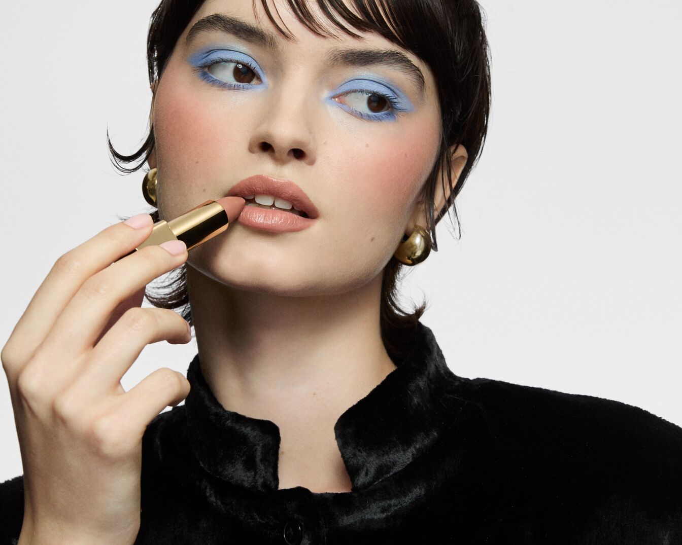 Eine Frau mit blauem Lidschatten und rosa Rouge trägt einen nudefarbenen Lippenstift aus einer goldenen Hülse auf.