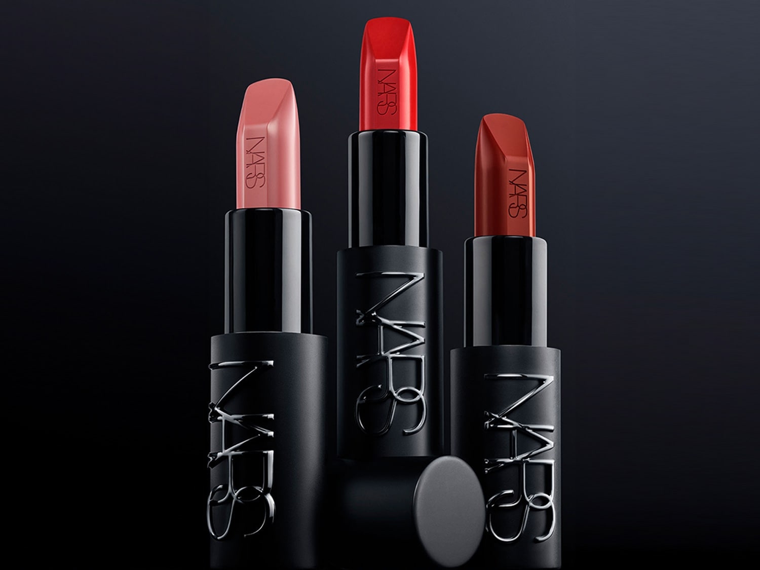 Tres barras de labios NARS en tonos rosa, rojo brillante y terracota, con envases negros que muestran el logo en plateado, sobre un fondo oscuro.