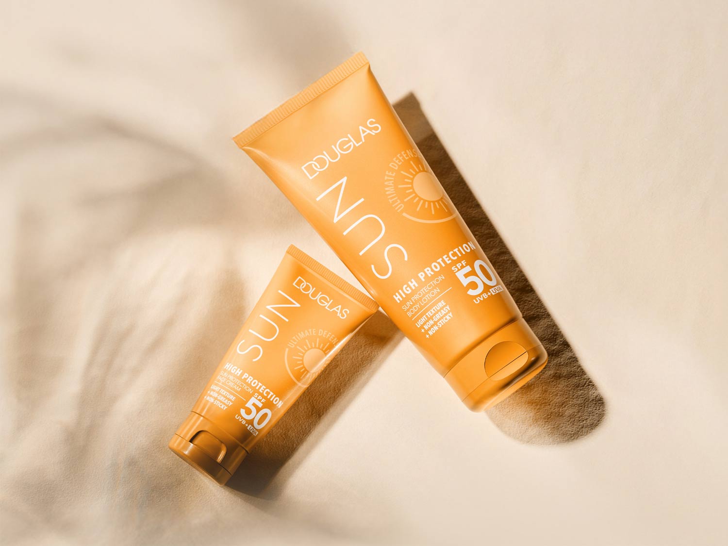 Deux tubes orange de crème solaire Douglas Sun Ultimate Defense SPF 50, offrant une haute protection avec une texture légère, non grasse et non collante, protégeant contre les rayons UVB et UVA.