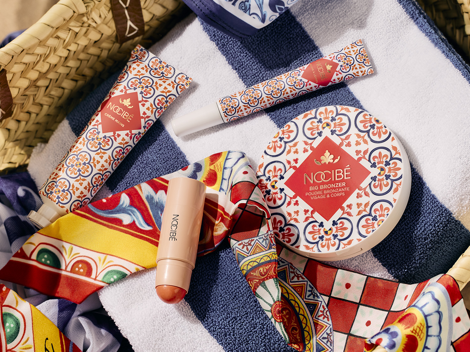 Dans un panier en osier, découvrez la crème pour les mains Nocibé, le soin pour les lèvres, le big bronzer visage et corps, et un stick rosé, le tout sur une serviette rayée bleu et blanche.