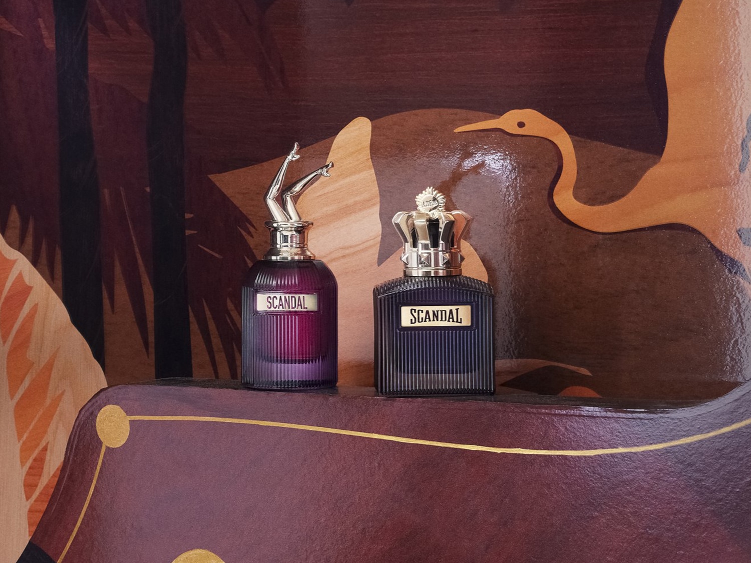 Két Scandal parfümös üveg látható: az egyik lila, a másik sötétkék, mindkettő egyedi kupakkal rendelkezik, az egyik egy ezüst színű koronával, a másik pedig egy pár ezüst színű lábbal.