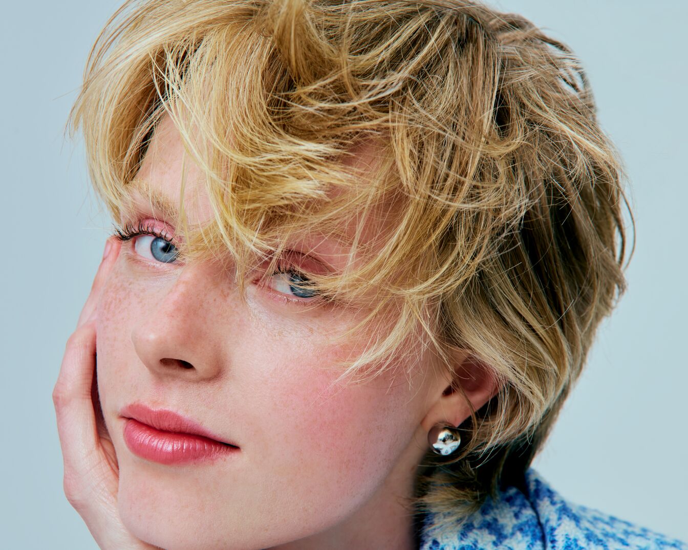 Close-up van een vrouw met blond haar, blauwe ogen en een roze make-up look, haar hand rustend op haar gezicht, met een zilveren oorbel.