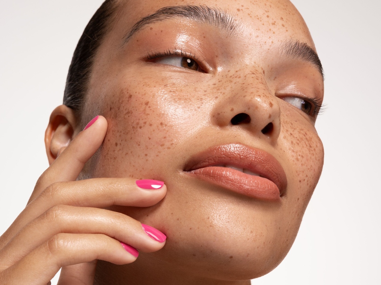 Portret kobiety z piegami, błyszczącą skórą i różowym manicure, która delikatnie dotyka swojej twarzy.