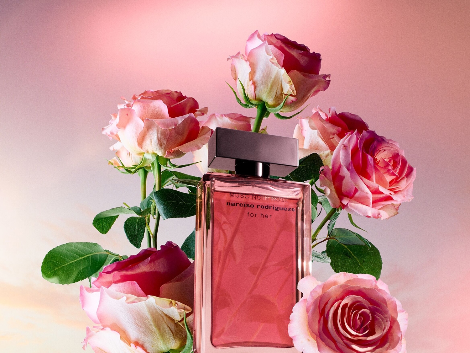 Ein rechteckiger, rosa Narciso Rodriguez "for her" Parfümflakon steht inmitten eines Bouquets aus rosa Rosen.