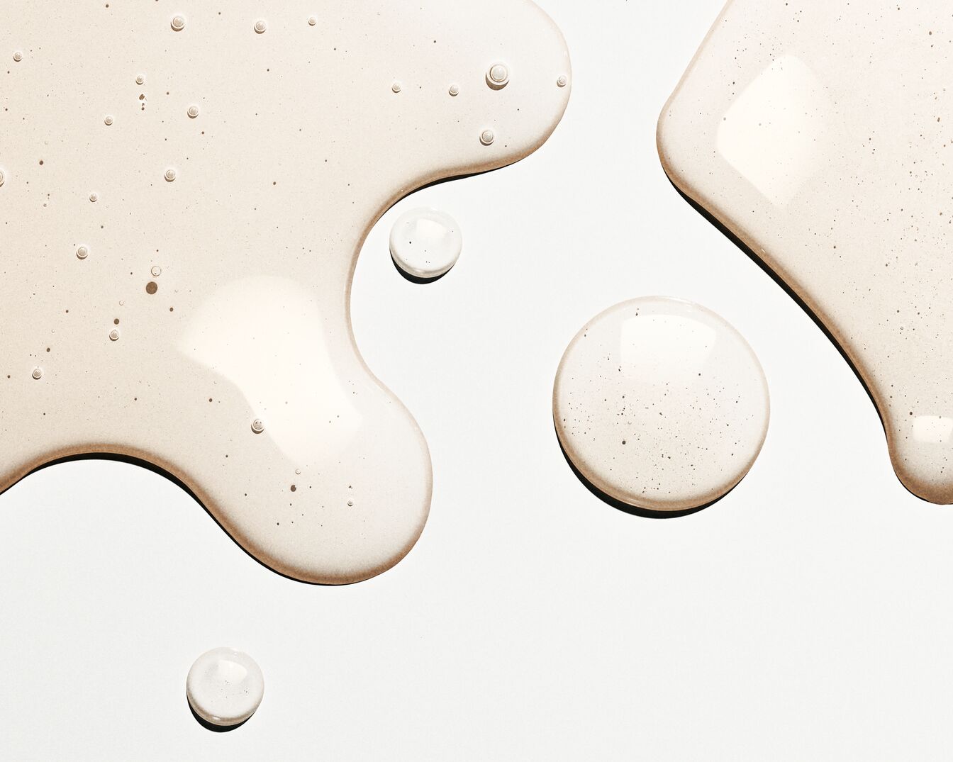 Nahaufnahme von transparenten, beigen Flüssigkeitstropfen und -pfützen mit kleinen Luftbläschen auf einer glatten, weißen Oberfläche.