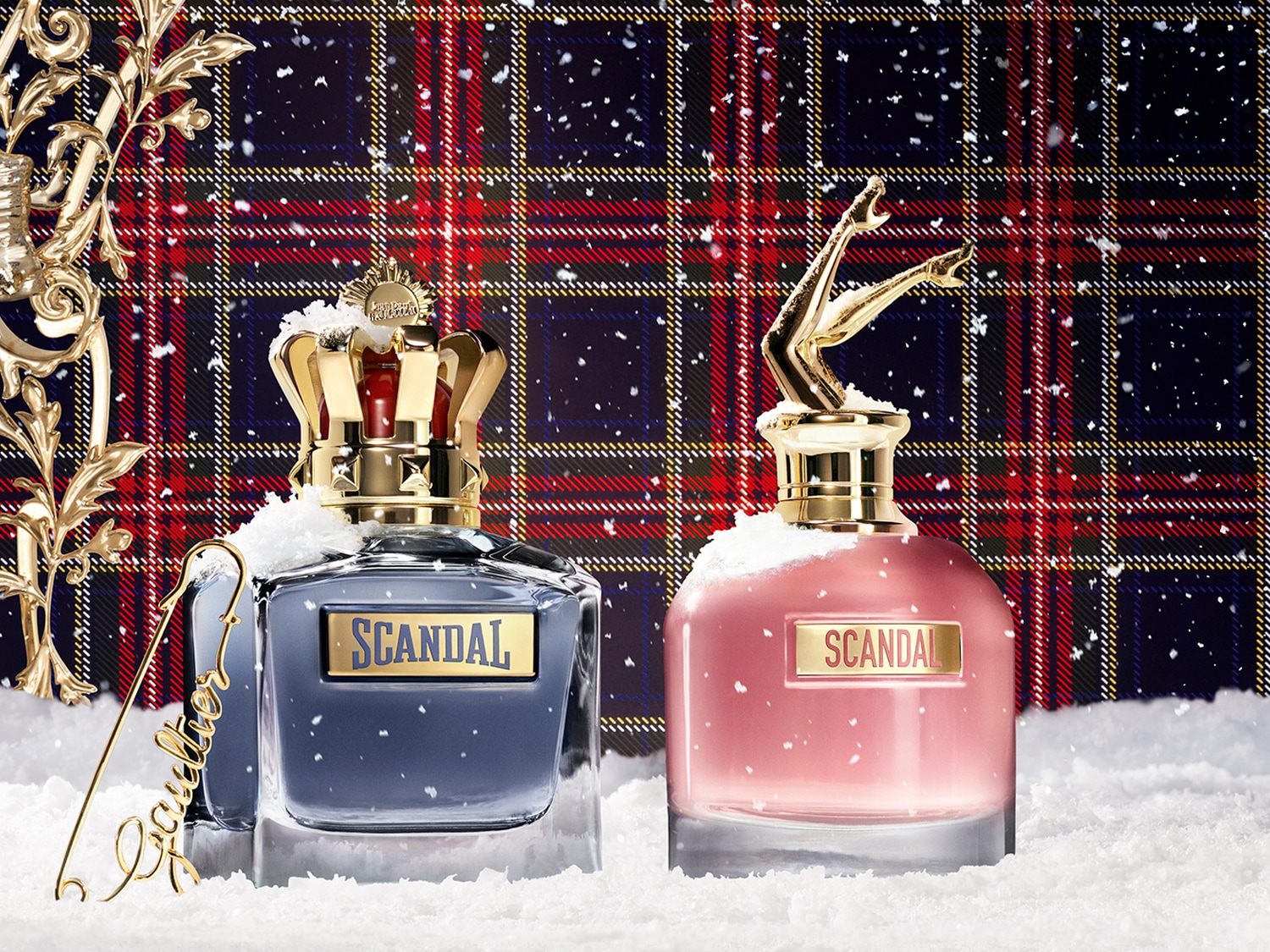 Dva flakóny vôní Scandal od Jean Paul Gaultier s viečkami v tvare korunky a nôh sú zasadené do zasneženej zimnej krajiny pred kockovaným pozadím.