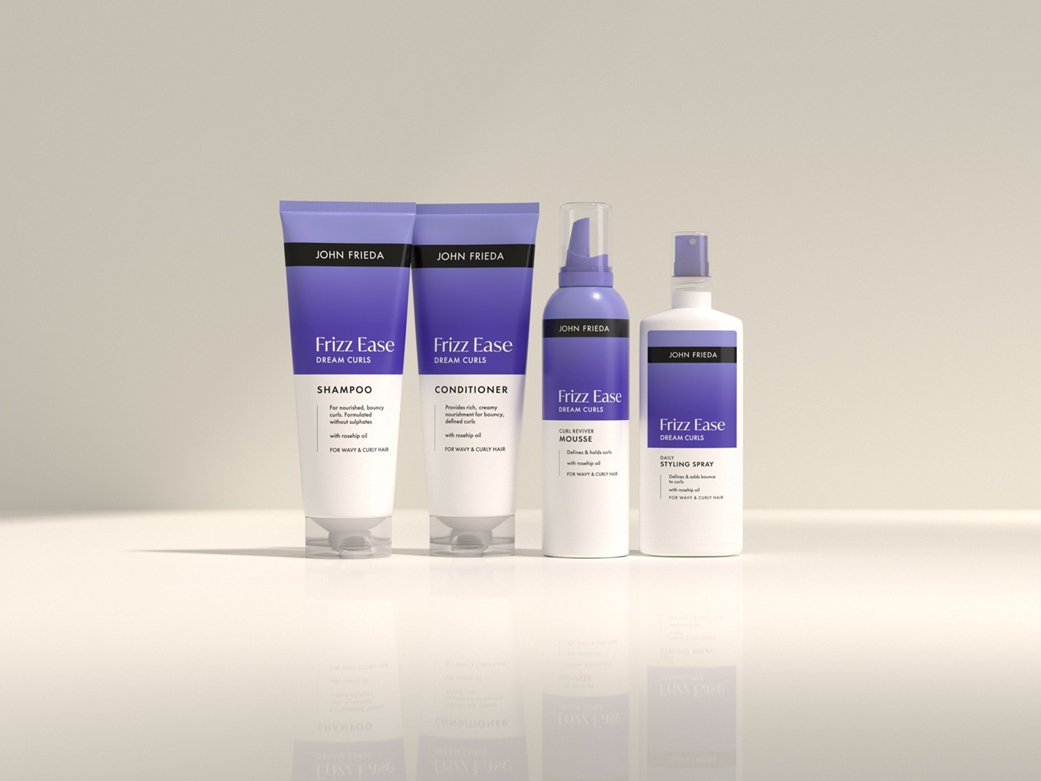 Čtyři produkty John Frieda Frizz Ease Dream Curls: šampon a kondicionér v bílých tubách s fialovými akcenty, pěna a stylingový sprej v bílých lahvích s fialovými detaily.