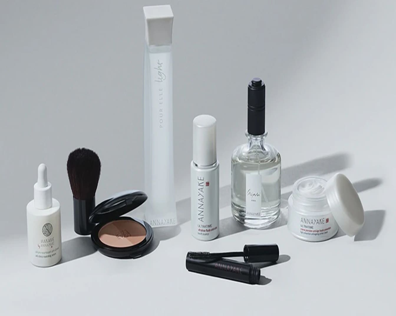 Eine Sammlung von ANNAYAKE-Produkten, darunter Seren, Puder, Parfüm und Mascara, die in eleganten weißen und schwarzen Verpackungen präsentiert werden.