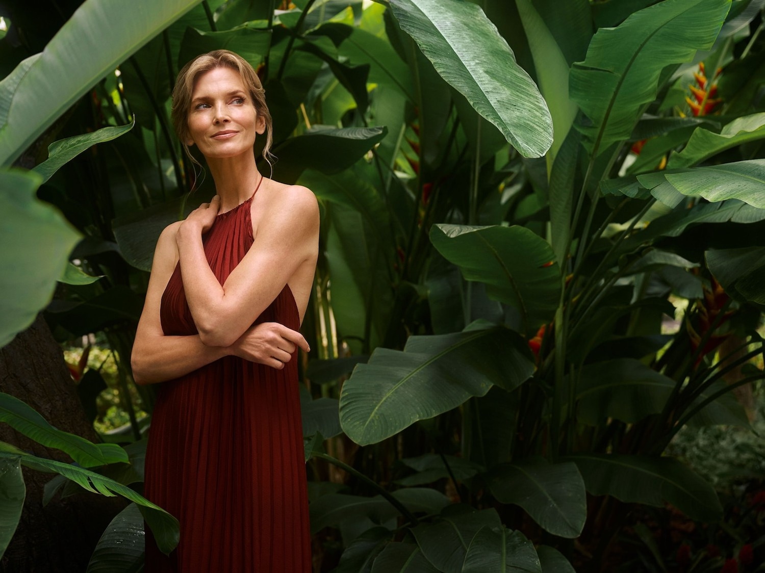 Eine Frau mit blonden Haaren und einem roten Kleid steht inmitten üppiger grüner Pflanzen und strahlt eine natürliche Eleganz aus.