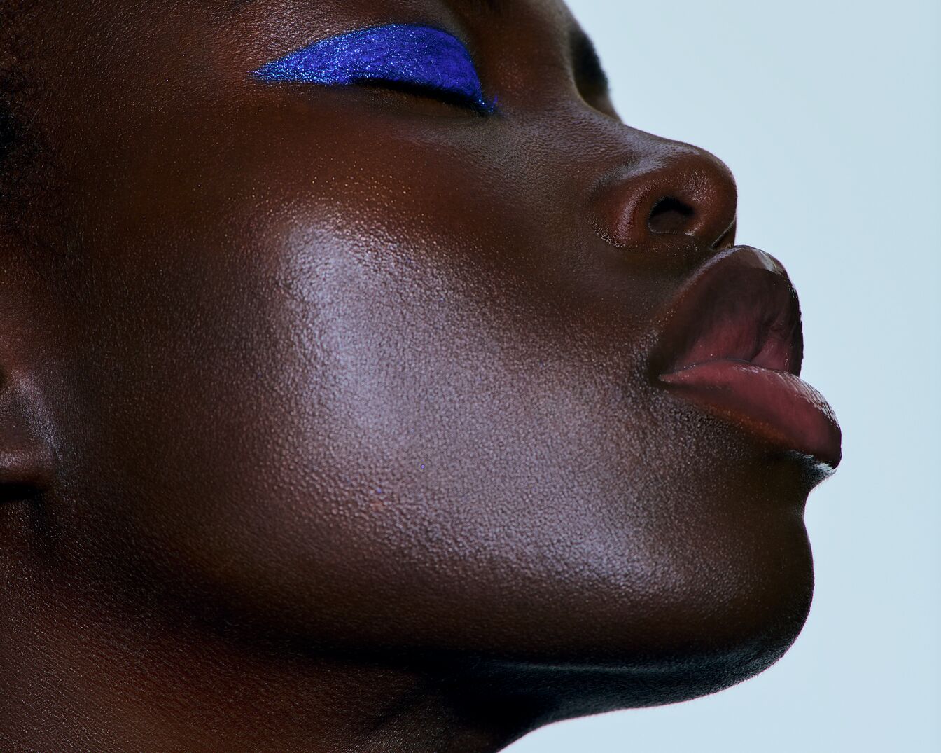 Nahaufnahme eines dunklen Hauttons mit blauem, glitzerndem Eyeliner und glänzenden Lippen, die einen modernen und auffälligen Look kreieren.