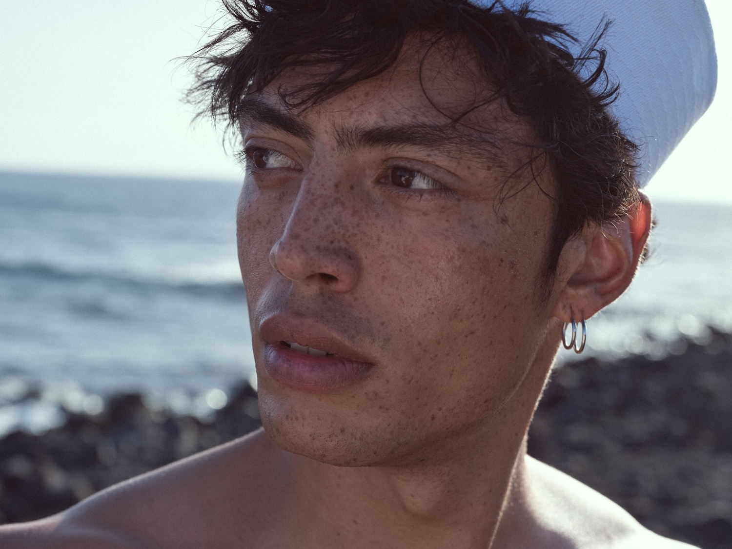Nahaufnahme eines Mannes mit Sommersprossen, der eine weiße Mütze und silberne Ohrringe trägt und zum Meer hinausschaut.