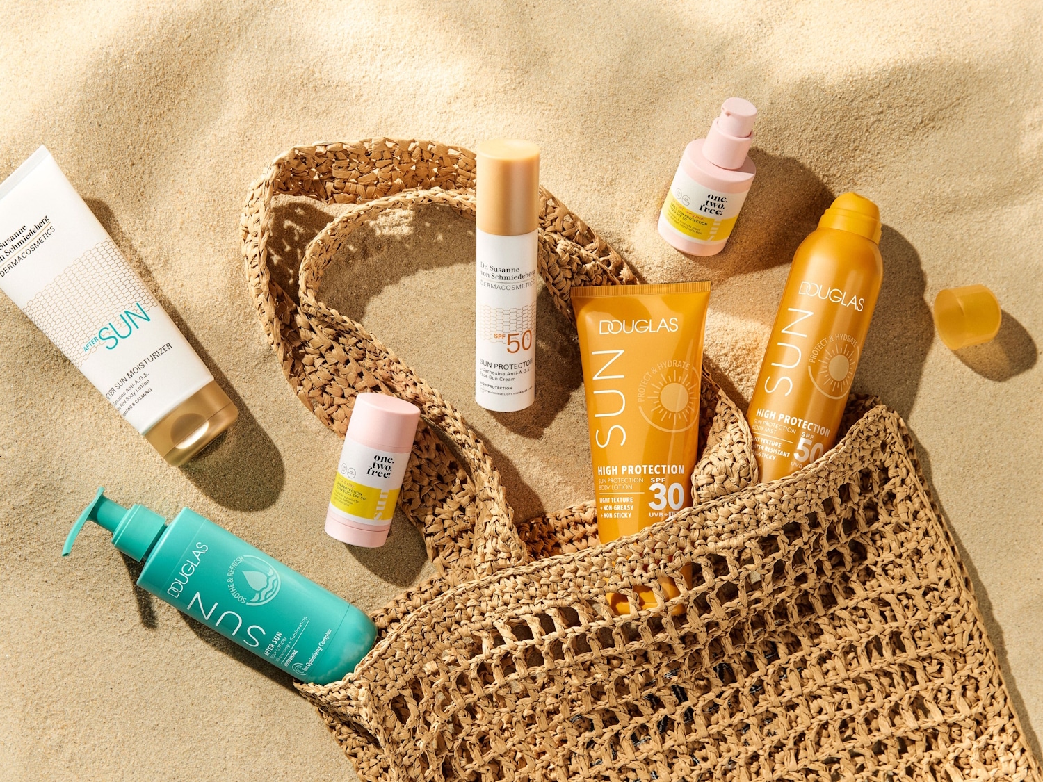 Verschiedene Sonnenschutzprodukte wie Lotionen und Sprays von Marken wie Douglas und Dr. Susanne von Schmiedeberg liegen auf einem Sandstrand neben einer Strandtasche.