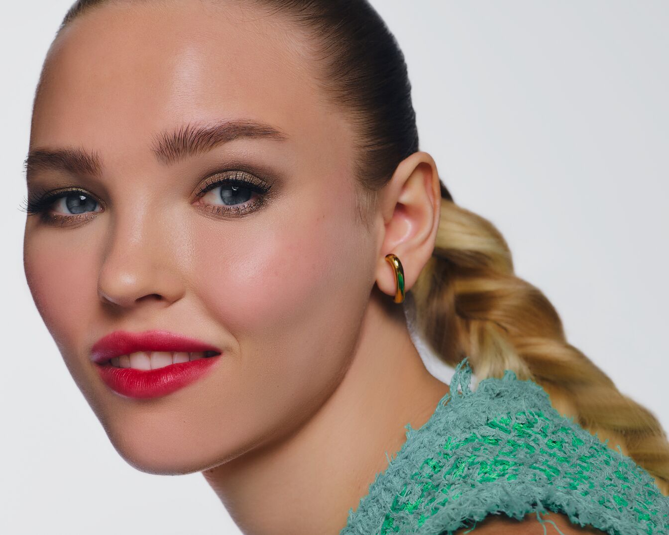 Nahaufnahme einer Frau mit strahlend rotem Lippenstift, goldenem Lidschatten und einem grünen Oberteil, die einen eleganten, geflochtenen Pferdeschwanz und goldene Ohrringe trägt.