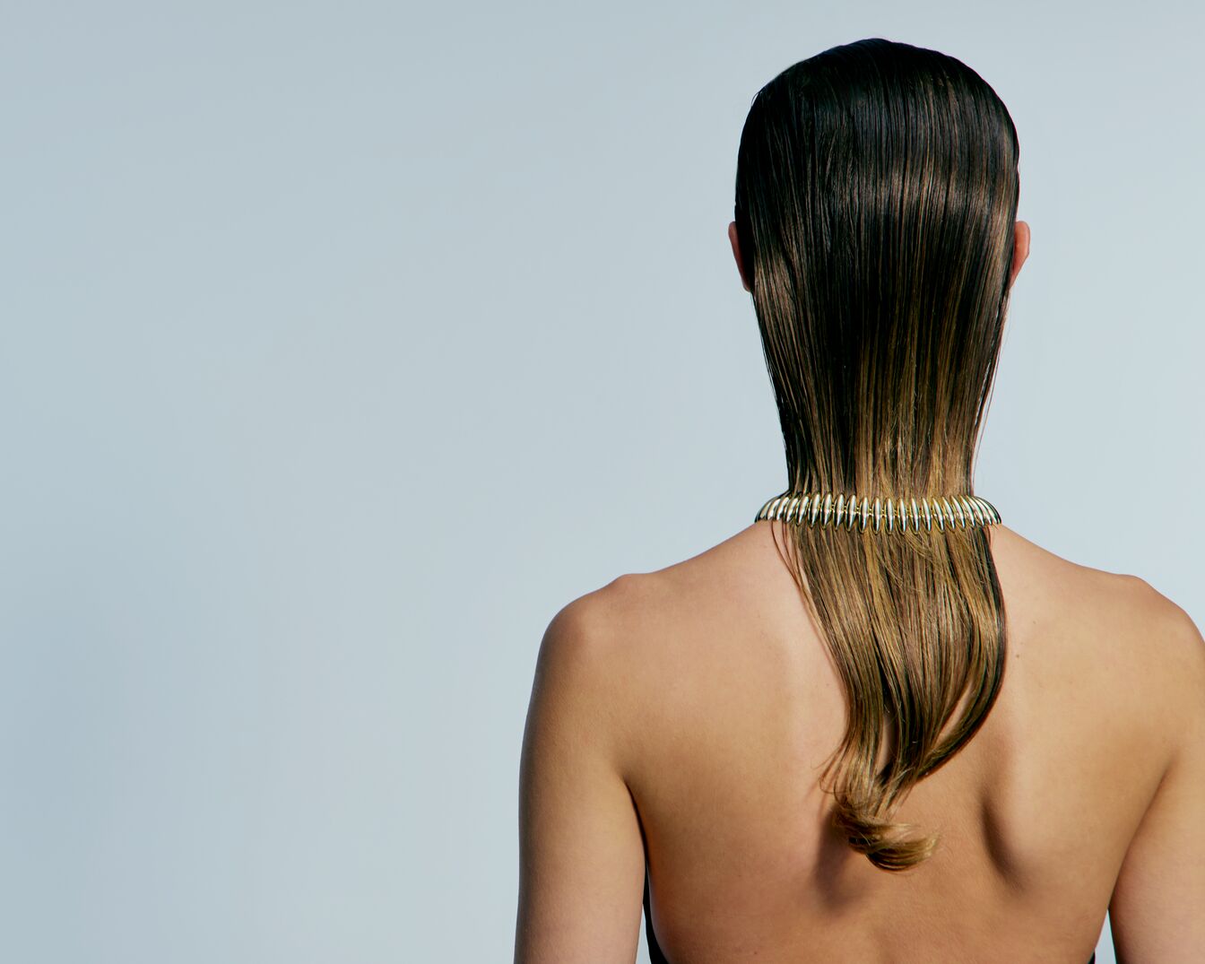 Een vrouw met halflang, bruin haar dat nat achterover is gekamd, draagt een gouden halsketting die op een veer lijkt.