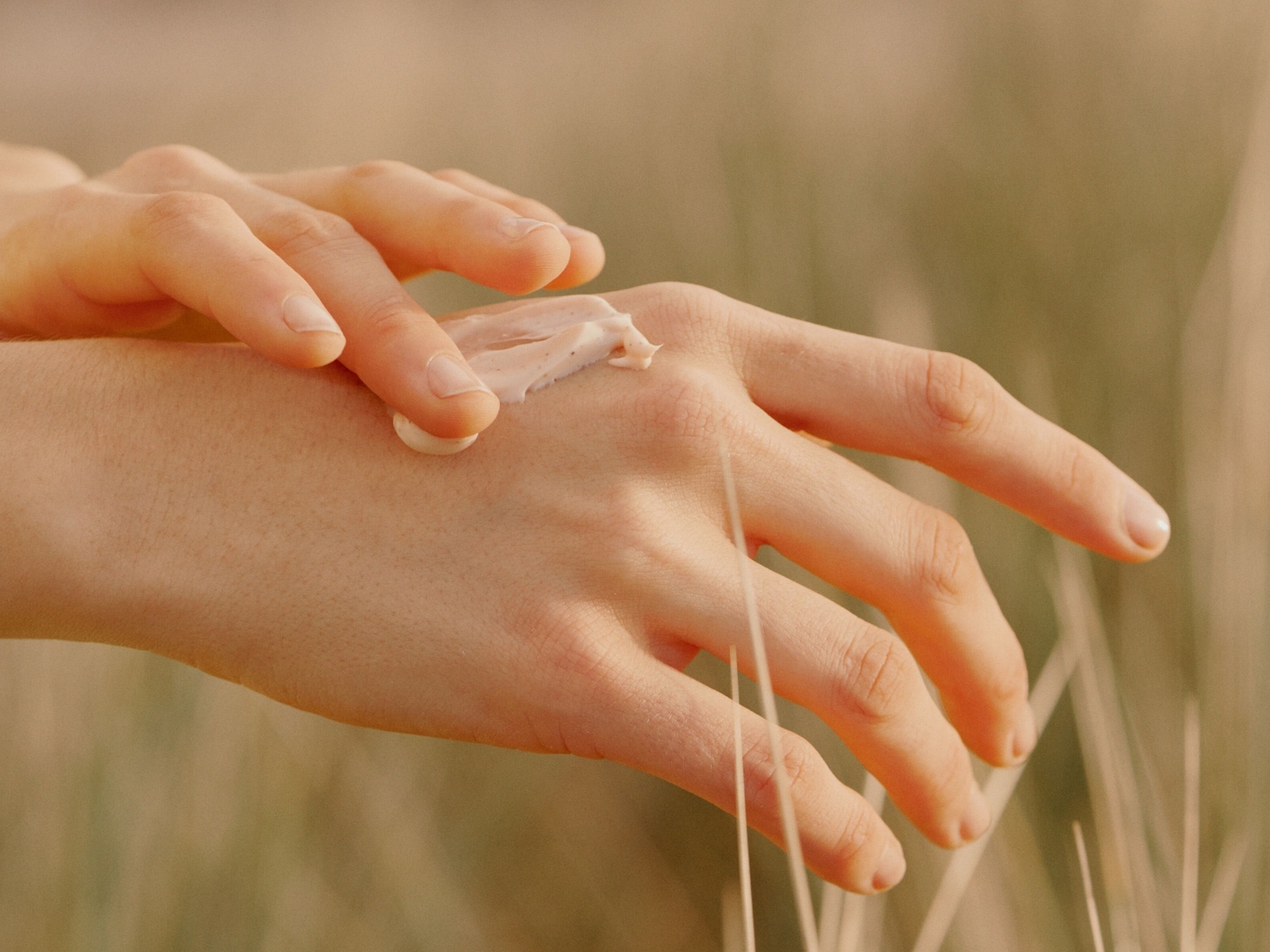 Nahaufnahme von zwei Händen, wobei eine Hand eine beigefarbene Creme auf die andere aufträgt, um die Haut mit Feuchtigkeit zu versorgen.