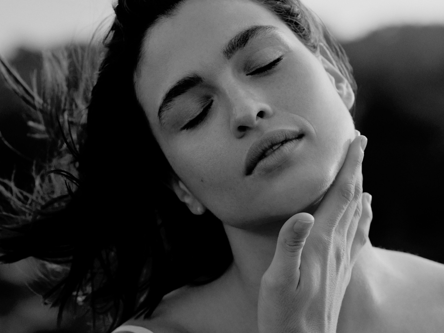 Een zwart-wit close-up van een vrouw met gesloten ogen, die zachtjes haar gezicht en hals aanraakt, wat een moment van rust en zelfzorg suggereert.